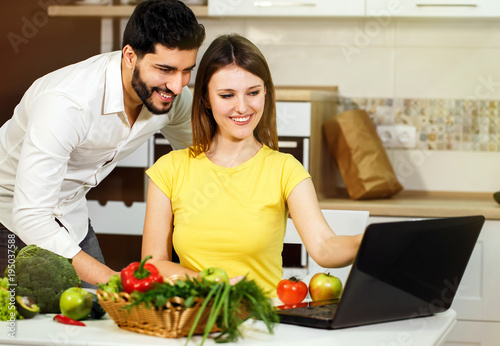 Lovely family couple spending evening together, smiling woman slicing cucumber, happy man standing nearby, using laptop and having pleasant conversation at the table with fresh green vegetables and