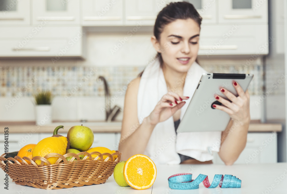 Attractive brunette using tablet in kitchen, after doing exercise while sitting at the table, light brown basket with fruits and meter in the foreground Attractive brunette using tablet in kitchen, after doing exercise while sitting at the table, light brown basket with fruits and meter in the foreground