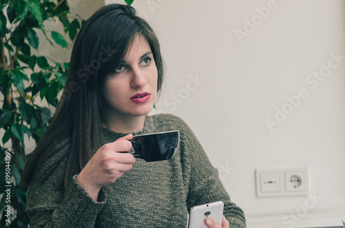 beautiful girl drinking tea and using a mobile phone while sitting in a cafe in front of a large window