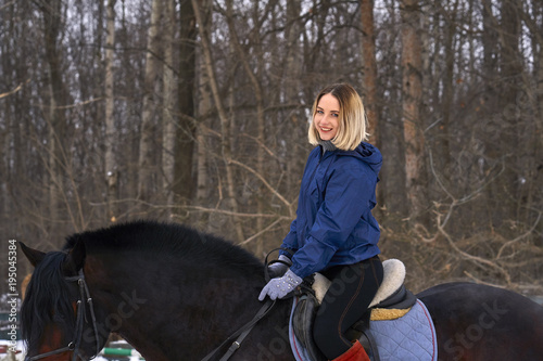 A young girl with white hair learns to ride a horse. The girl recently started to practice equestrianism. The girl is afraid of riding a horse quickly. A cloudy winter day..