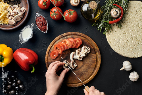Female hand cut mushrooms and tomatoes on wooden board on kitchen table, around lie ingredients for pizza: vegetables, cheese and spices.