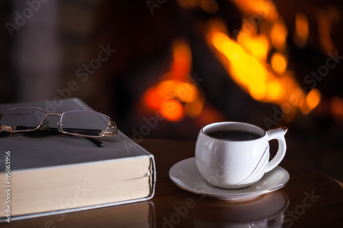 White cup of tea or coffee, glasses and old book near fireplace on wooden table. Winter and Christmas holiday concept