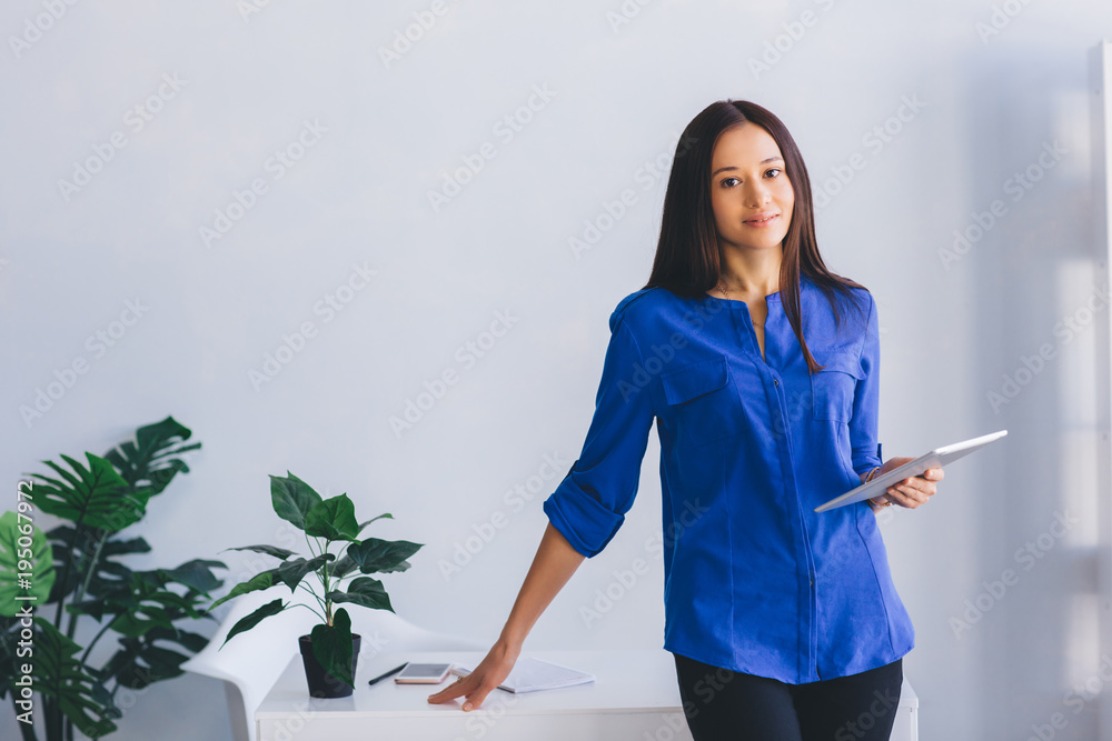 Modern, confident young woman in stylish clothes holding digital tablet, smiling while standing near her working place at office. Modern, confident young woman in stylish clothes holding digital tablet, smiling while standing near her working place at office.