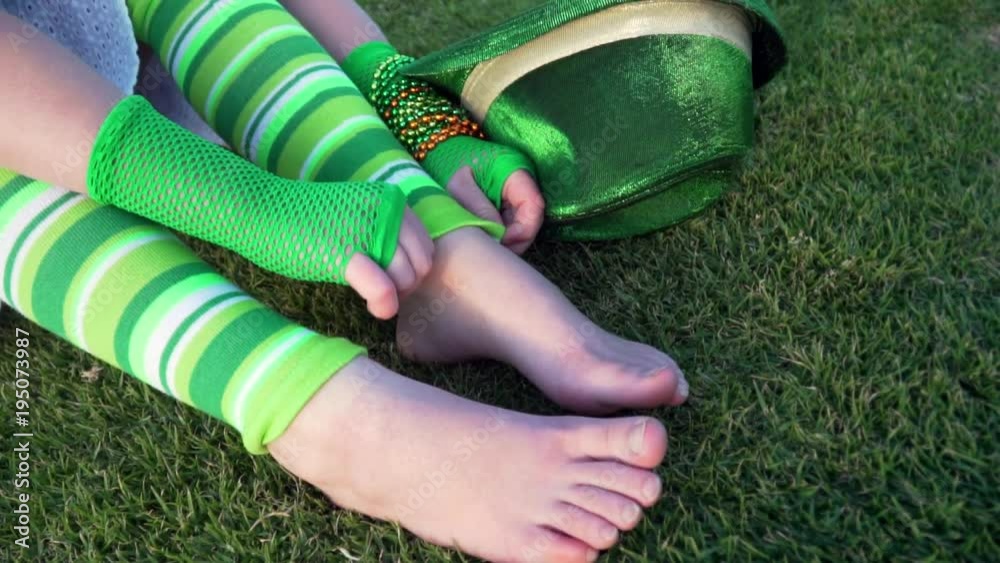 Slow motion. Girl in funny green striped leg warmers fixing it. Saint Patrick's day. Green gloves, hat, bracelets on the wrist. Birthday party. Close up ancle. Vert gants filets. Grass, park. Carnival