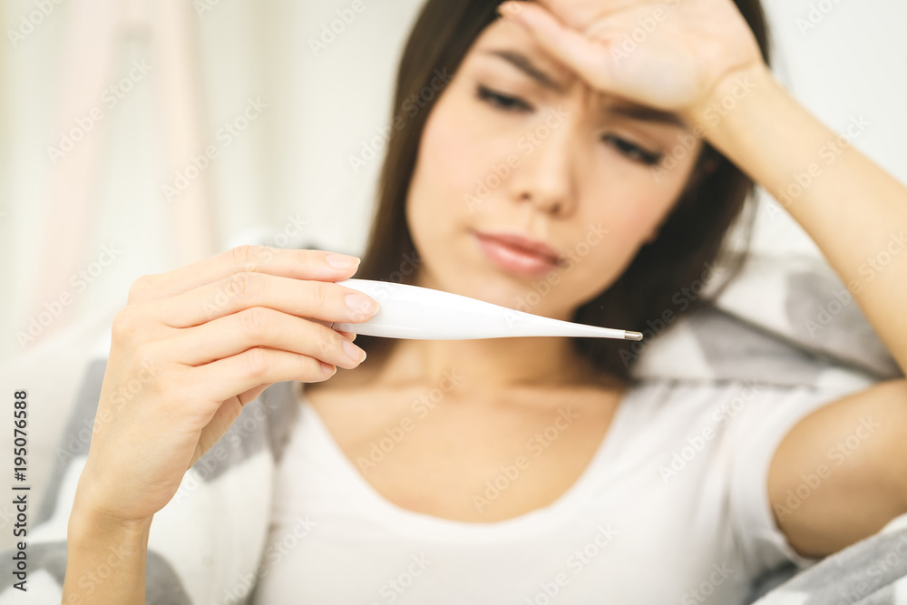 Woman with flu virus lying in bed, she is measuring her temperature with a thermometer and touching her forehead. Close-Up Woman with flu virus lying in bed, she is measuring her temperature with a thermometer and touching her forehead. Close-Up