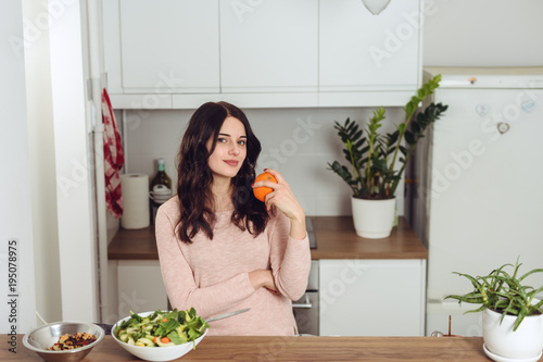 Smiling young woman in the kitchen near desk and holds a fruit in his hand. Healthy life concept