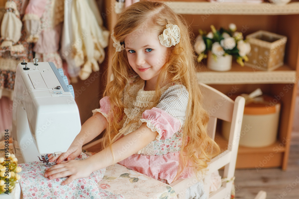 a small blonde girl with long hair smiling beautiful baby girl 6 years in a  retro dress sew on a sewing machine is a beautiful  style of baby  chic Stock Photo |