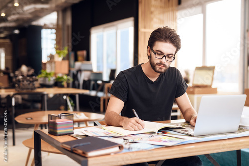 Freelancer bearded man taking notes at laptop sitting at desk. photo