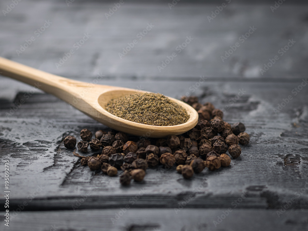 A wooden spoon with ground black pepper on a pile of whole pepper on a black wooden table. A wooden spoon with ground black pepper on a pile of whole pepper on a black wooden table.