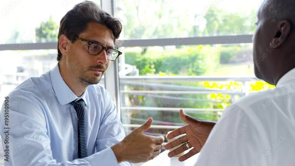 Two Businessmen Shaking Hands Around Table In Modern Office Two Businessmen Shaking Hands Around Table In Modern Office