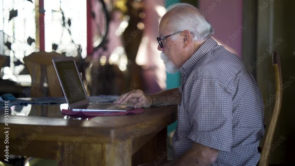 Backlit elderly man with white beard working on laptop computer at a table. Backlit elderly man with white beard working on laptop computer at a table.