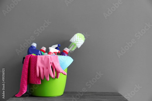 Bucket with cleaning supplies and tools on table near gray wall