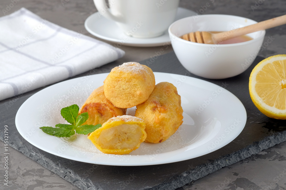 Bananas in batter and sprinkled with powdered sugar on a white plate on the gray slate background. The bowl with honey, lemon and cup of tea near it. Bananas in batter and sprinkled with powdered sugar on a white plate on the gray slate background. The bowl with honey, lemon and cup of tea near it.
