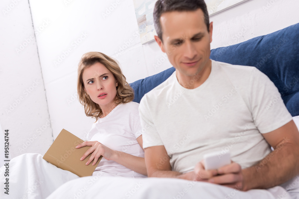 selective focus of man using smartphone while wife reading book in bed selective focus of man using smartphone while wife reading book in bed
