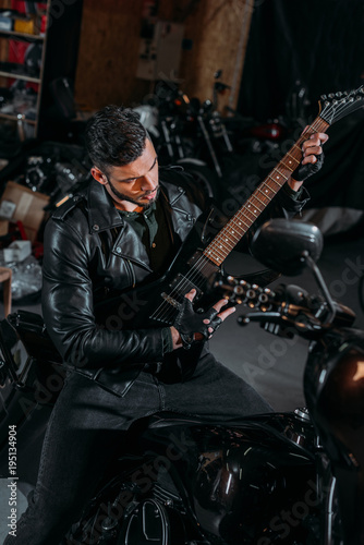 handsome young man playing guitar while sitting on bike at garage