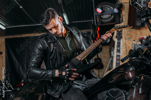handsome young man playing guitar while sitting on bike at garage