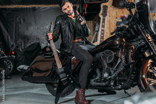 stylish young man in leather jacket sitting on bike with electric guitar at garage