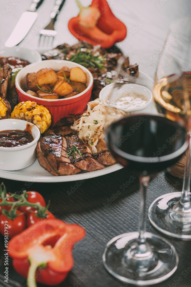plate with sauces, roasted potatoes, grilled meat with vegetables and glasses of wine on table