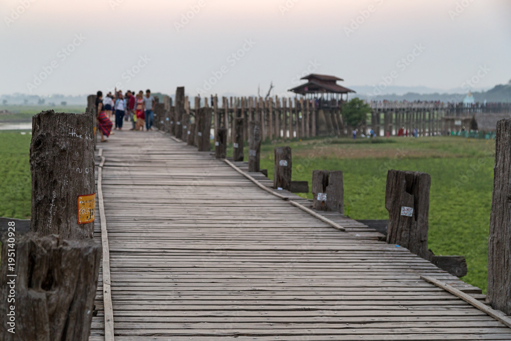 Obraz premium Some people at the U Bein Bridge at Amarapura near Mandalay in Myanmar (Burma) at dusk. It's the oldest and longest teakwood bridge in the world. Focus on the foreground.