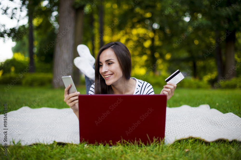 Attractive smiling young woman with a bank card and phone orders goods lying on a plaid in the park. Online shopping Attractive smiling young woman with a bank card and phone orders goods lying on a plaid in the park. Online shopping