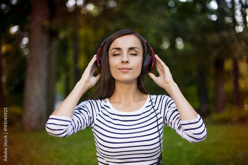 Concentrated beautiful young woman with closed eyes listening to music on the big headphones on a sunset in the park