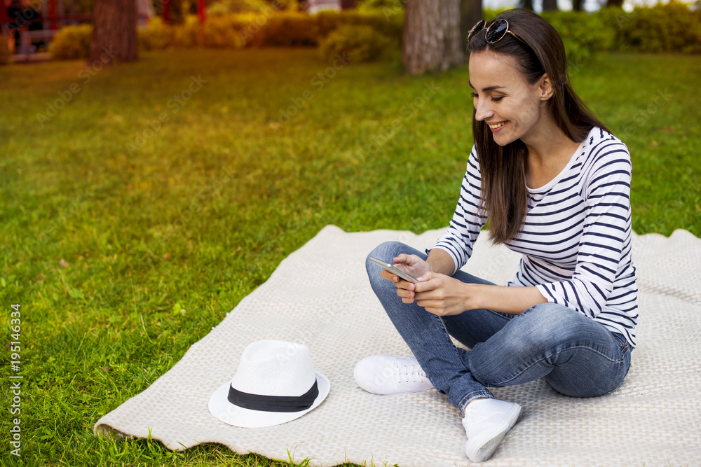 Beautiful smiling modern young girl student in casual clothes sitting on a plaid in the park with a phone Beautiful smiling modern young girl student in casual clothes sitting on a plaid in the park with a phone