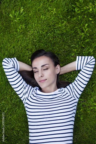 A beautiful smiling modern young woman with gorgeous hair and closed eyes lies on a green lawn. View from above. Concept of sleep and rest