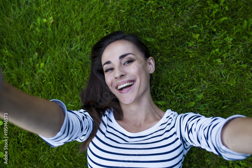 An attractive smiling modern young woman with chic hair and in casual clothes lies on the green grass. View from above