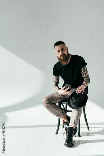 handsome bearded tattooed man sitting on chair with smartphone and hat on white