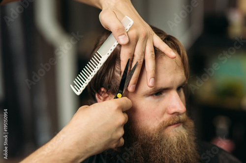 The hands of young barber making haircut to attractive bearded man in barbershop