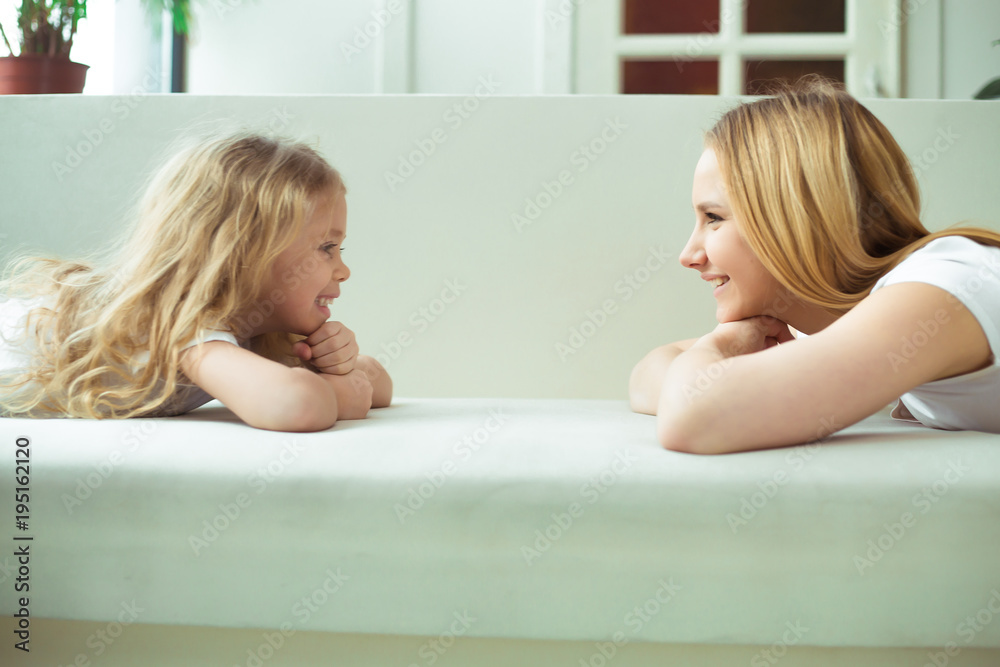 Moms and daughters. Young beautiful woman and a little blond girl lie on a white sofa face to face and look at each other.