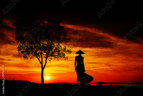 Silhouette of a young and fit woman with an Asian conical hat on the beach at sunset