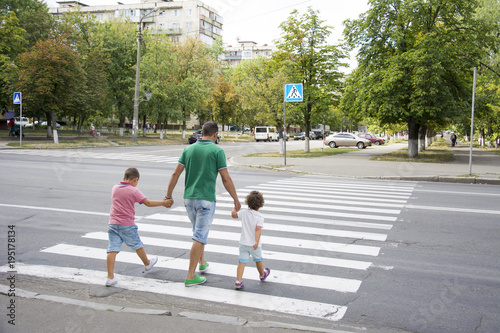 In the summer on the street at the pedestrian crossing father and son and daughter cross the road. Dad keeps the children by the hands.