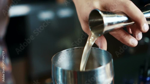 Close up of yellow liquer is being poured to steel shaker from measure glass in modern bar, slow motion. Barmen is inclining the cup by his hand and delicious drink is falling to the equipment for