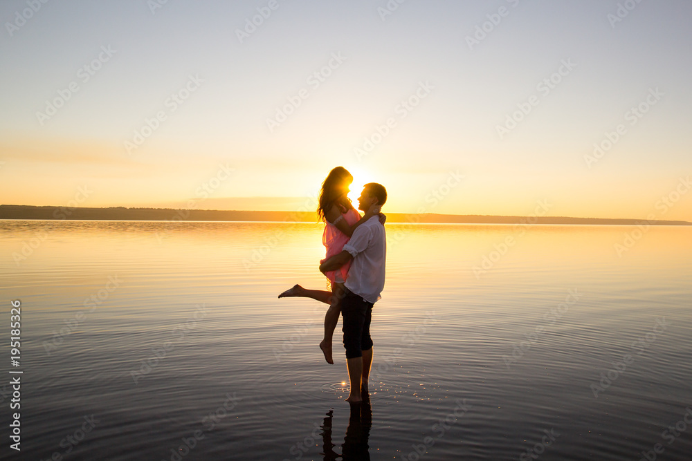 Fototapeta premium Young couple is embracing in the water on summer beach. Sunset over the sea.Two silhouettes against the sun. Just married couple hugging. Romantic love story. Man and woman in holiday honeymoon trip.