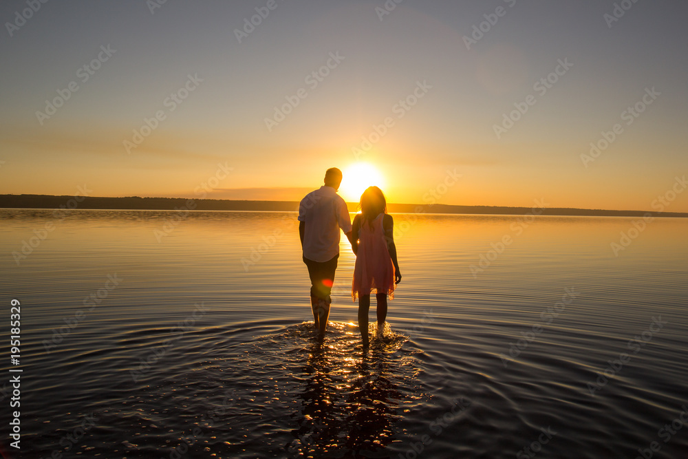 Young couple is walking in the water on summer beach. Sunset over the sea.Two silhouettes against the sun. Just married couple hugging. Romantic love story. Man and woman in holiday honeymoon trip.