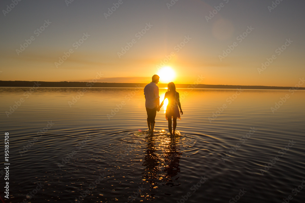Fototapeta premium Young couple is walking in the water on summer beach. Sunset over the sea.Two silhouettes against the sun. Just married couple hugging. Romantic love story. Man and woman in holiday honeymoon trip.