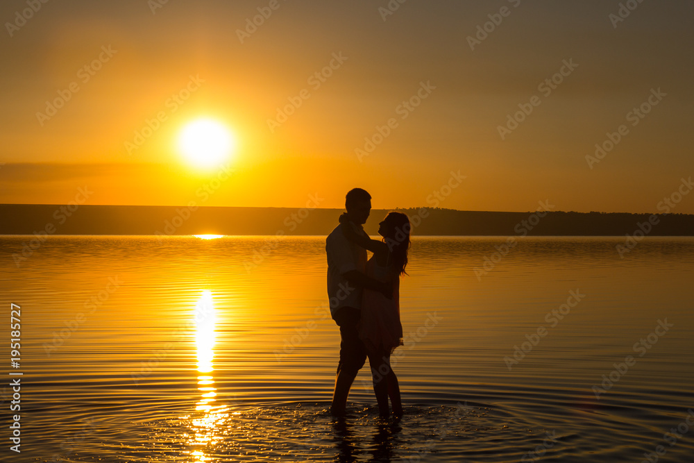 Young couple is embracing in the water on summer beach. Sunset over the sea.Two silhouettes against the sun. Just married couple hugging. Romantic love story. Man and woman in holiday honeymoon trip.