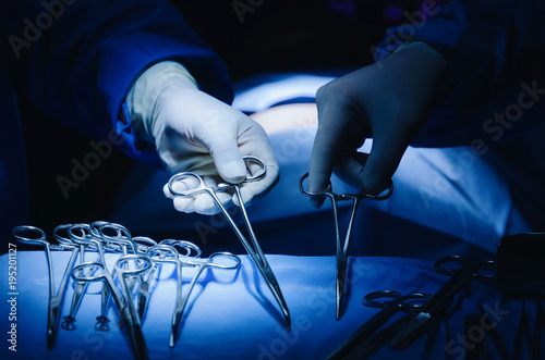 close up surgeons hand taking scissors, forceps and surgical instruments on table for operation with colleagues performing work in operation room at hospital, emergency case, surgery, medical concept