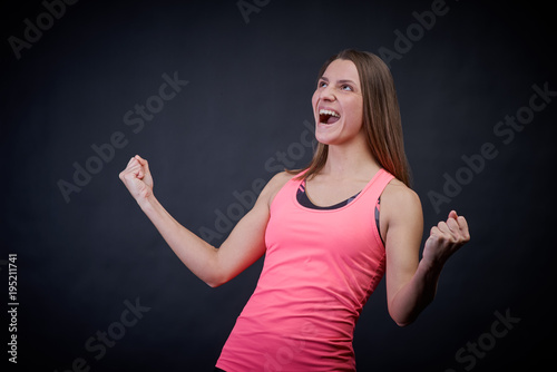 Stunning woman in sports uniform on an isolated black background keeps her fists from happiness, showing victory, looking sideways and upstairs, the athlete enjoys victory and first place