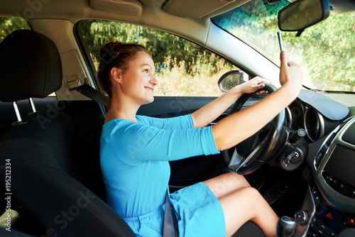 Happy young woman holding mobile phone and taking photos while driving a car. Smiling girl taking selfie picture with cellphone camera outdoors in car. Holidays and tourism concept