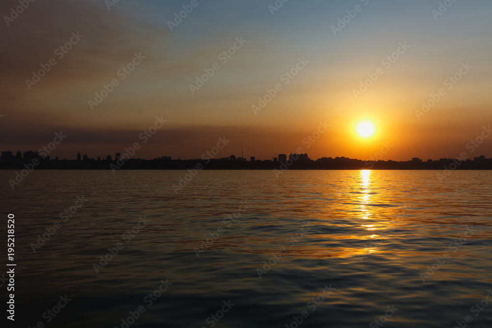Beautiful sea landscape. Bright warm sunset in the sea near the coastline with the city's silhouettes. Sun hides behind the house. Colors of the sky are reflected in the water