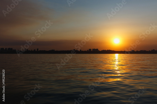 Beautiful sea landscape. Bright warm sunset in the sea near the coastline with the city s silhouettes. Sun hides behind the house. Colors of the sky are reflected in the water