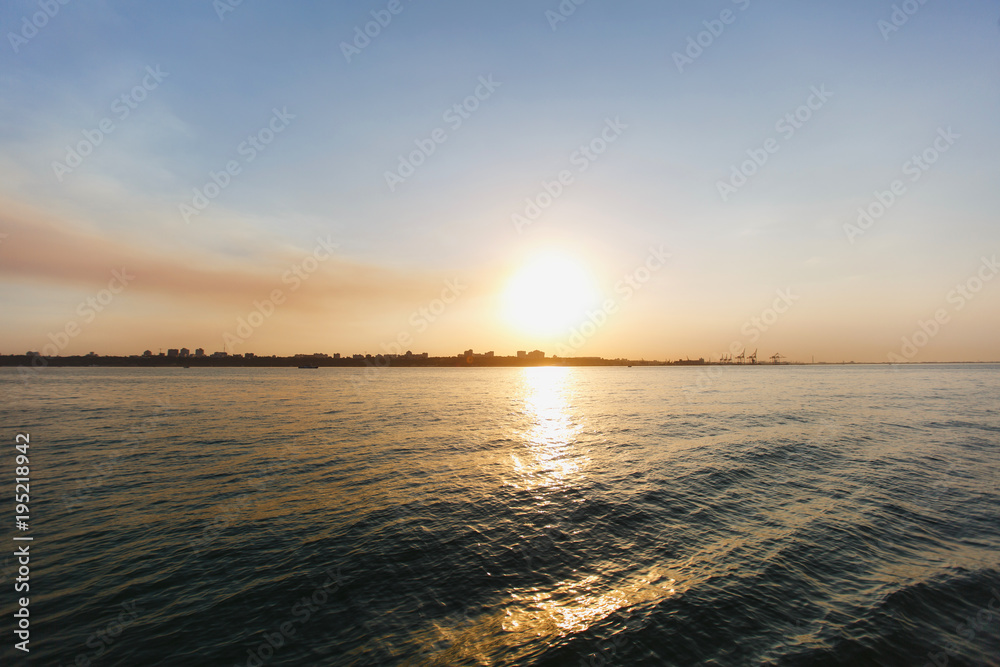 Naklejka premium Beautiful sea landscape. Bright warm sunset in the sea near the coastline with the city's silhouettes. Sun hides behind the house. Colors of the sky are reflected in the water