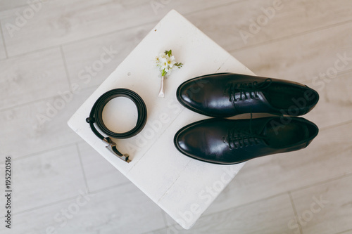 Close up Beautiful elegant wedding polished black shoes with shoelaces, belt and flower boutonniere for the groom on a chair on background a light wooden floor. Wedding wear, accessories photo