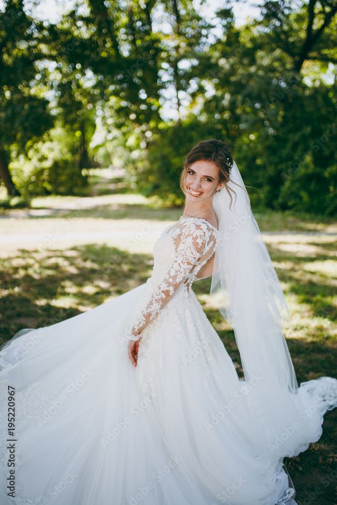 Beautiful wedding photosession. The smiling bride in a white lace dress with a plume, long sleeves, hairpin in hair, white veil turns around in a large green garden on weathery sunny day