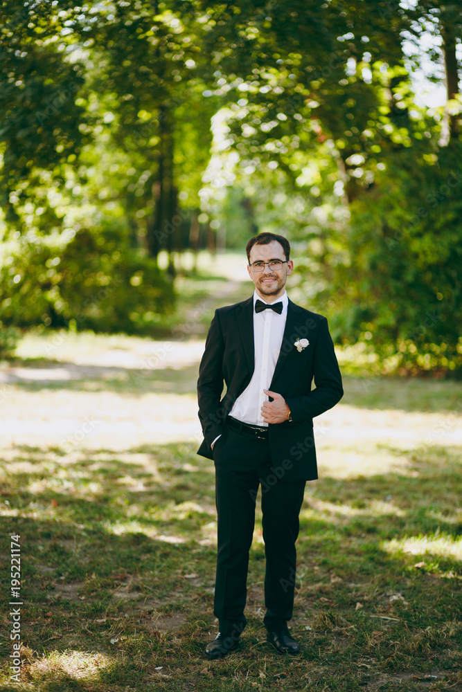 Beautiful wedding photosession. The young smiling unshaved groom in a black suit, a white shirt, a bow tie and glasses with a boutonniere of flowers in a large green garden on weathery sunny day Beautiful wedding photosession. The young smiling unshaved groom in a black suit, a white shirt, a bow tie and glasses with a boutonniere of flowers in a large green garden on weathery sunny day
