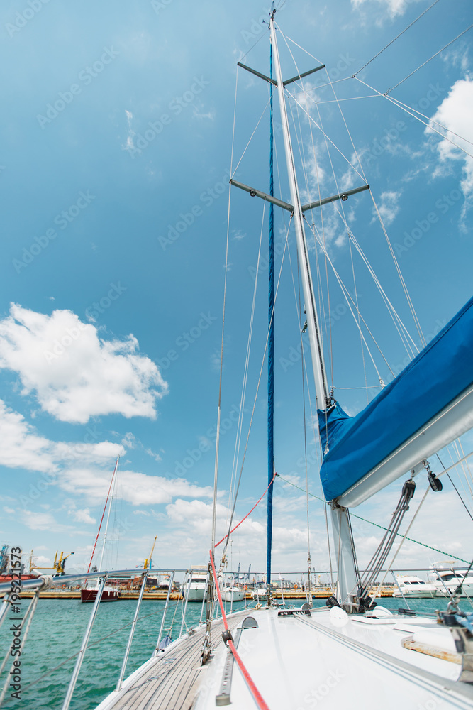 Sailing boat deck on a white yacht with lowered sails and teak wooden deck, metal railing, set of red and blue ropes in the sea near the port in the background blue sky with clouds in sunny day
