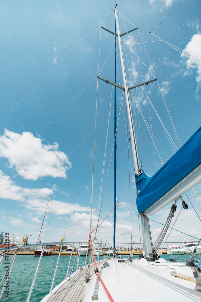 Sailing boat deck on a white yacht with lowered sails and teak wooden deck, metal railing, set of red and blue ropes in the sea near the port in the background blue sky with clouds in sunny day Sailing boat deck on a white yacht with lowered sails and teak wooden deck, metal railing, set of red and blue ropes in the sea near the port in the background blue sky with clouds in sunny day