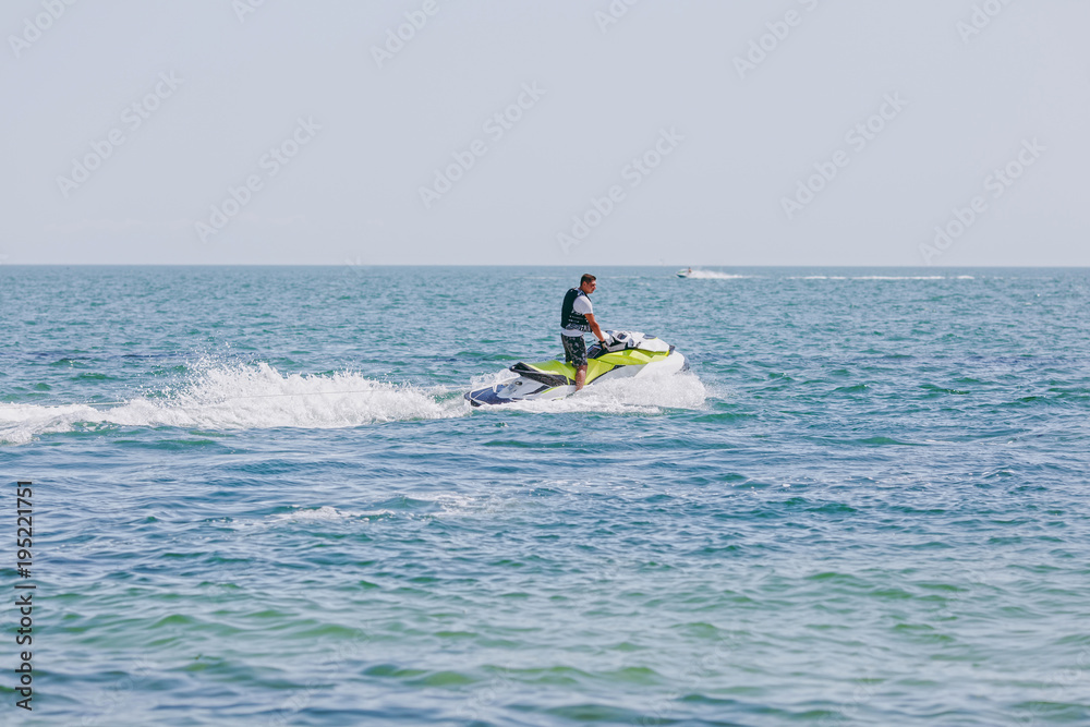 Young man in shorts, t-shirt and lifejacket riding a white and yellow water scooter on the blue high seas, forming large waves, splashes and foam on the background clear sky